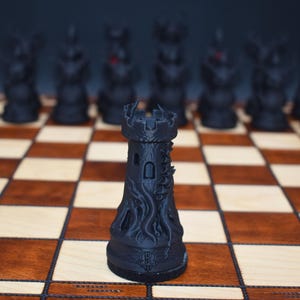 May include: A black chess rook piece with a detailed, textured design, standing on a wooden chessboard with brown and beige squares. Other black chess pieces are blurred in the background.