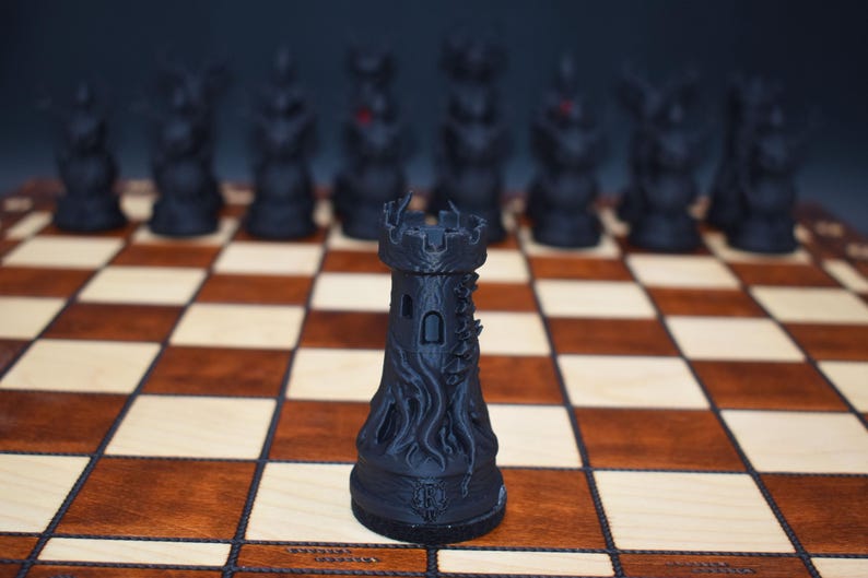 May include: A black chess rook piece with a detailed, textured design, standing on a wooden chessboard with brown and beige squares. Other black chess pieces are blurred in the background.