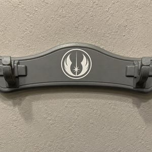 K&ouml;nnte beinhalten: Eine graue Lichtschwert-Wandhalterung mit einem wei&szlig;en Emblem mit Stern und Fl&uuml;geln. Die Halterung hat zwei Clips zum Halten von Lichtschwertern und ist an einer strukturierten Wand montiert.