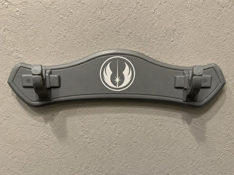 K&ouml;nnte beinhalten: Eine graue Lichtschwert-Wandhalterung mit einem wei&szlig;en Emblem mit Stern und Fl&uuml;geln. Die Halterung hat zwei Clips zum Halten von Lichtschwertern und ist an einer strukturierten Wand montiert.