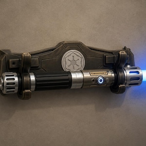 K&ouml;nnte beinhalten: Eine bronzefarbene Lichtschwert-Wandhalterung mit einem silbernen und schwarzen Lichtschwert. Das Lichtschwert strahlt einen hellblauen Strahl aus. Die Wandhalterung hat ein Symbol in der Mitte.