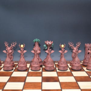 May include: A unique chess set with pieces designed to resemble trees and forest elements. The pieces are a reddish-brown color and include rooks, deer, and pieces with glowing or leafy tops. The set is displayed on a wooden chessboard.