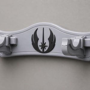 Puede incluir: Un soporte de pared gris claro con dos brazos de apoyo curvos y un símbolo de la Orden Jedi negro en el centro. El montaje horizontal tiene un acabado moldeado liso con un borde superior arqueado decorativo y una estructura sólida.