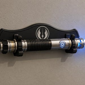 K&ouml;nnte beinhalten: Ein Lichtschwert mit silbernem und schwarzem Griff, das eine hellblaue Klinge aussendet. Das Lichtschwert ist auf einer schwarzen Wandplakette mit einem wei&szlig;en Emblem montiert. Es ist ein Requisit aus dem Star Wars-Franchise.