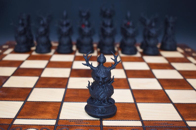 May include: A black chess set with intricately designed pieces on a wooden chessboard. The focal point is a detailed black chess piece with antlers, flames, and a skull-like face. The board has alternating light and dark squares.