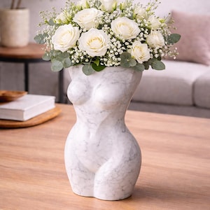 May include: A white marble vase in the shape of a female torso, filled with white roses, baby's breath, and greenery. The vase sits on a wooden table, with a blurred background of a living room.