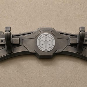 K&ouml;nnte beinhalten: Ein dunkelgrauer, an der Wand montierter Kleiderhaken mit zwei Haken und einem zentralen Emblem. Der Haken hat ein geometrisches Design mit einem erhabenen, kreisf&ouml;rmigen Emblem in der Mitte. Die Haken sind schwarz und rechteckig. Der Haken ist aus einem haltbaren Material.