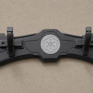 K&ouml;nnte beinhalten: Ein schwarzer, an der Wand montierter Displayst&auml;nder mit zwei Haken. Der St&auml;nder hat ein zentrales Emblem und ist f&uuml;r die Aufnahme eines Lichtschwerts konzipiert. Der St&auml;nder besteht aus einem haltbaren Material und ist f&uuml;r die Wandmontage konzipiert.