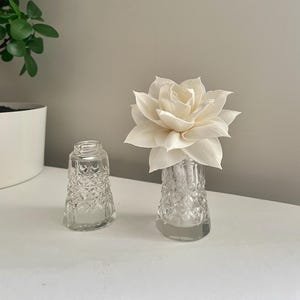 May include: Two clear glass salt and pepper shakers with a diamond pattern. One shaker is empty, and the other holds a large, white, paper flower. The objects are on a white surface.