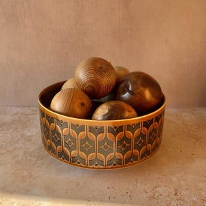 May include: A round ceramic bowl featuring a repeating dark green and orange geometric pattern on the exterior. The bowl is filled with several smooth, spherical wooden objects of varying brown tones and natural wood grain textures.