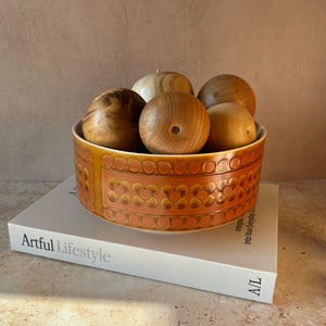 May include: A round ceramic bowl with a glossy orange and brown finish featuring a repeating embossed floral pattern. The bowl is filled with several smooth, light-coloured wooden spheres of varying grain patterns and sits atop a white book.