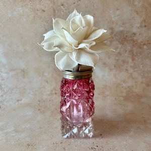 May include: A small, square-shaped glass bottle with a diamond-cut pattern that transitions from clear at the base to a deep pink at the top. The bottle is topped with a metallic cap and a large, cream-coloured, multi-petaled wooden flower.