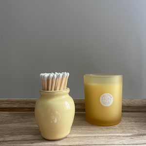 May include: A small, pale yellow ceramic match holder filled with wooden matches featuring white tips, positioned next to a frosted yellow glass candle jar with a circular white label containing text on the front surface.