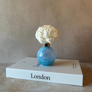 May include: A small, spherical blue and white marbled glass vase with a gold-toned neck, topped with a large, cream-coloured artificial peony flower. The vase rests on a white hardcover book with the word London printed on the front cover.