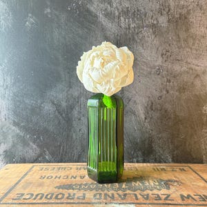 May include: A tall, hexagonal, emerald green glass vase with vertical ribbed detailing. A single, large, white peony flower with layered, ruffled petals is positioned inside the vase, extending upwards from the narrow opening at the top.