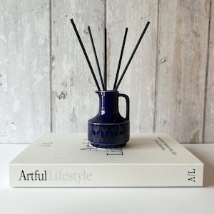 May include: A dark blue ceramic reed diffuser with a handle and a spout, featuring a repeating grey leaf-like pattern around the base. Five thin black reeds are inserted into the top of the vessel, which sits atop a white book titled Artful Lifestyle.