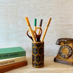 May include: A cylindrical ceramic pencil holder with a brown and black geometric pattern containing four pens and a pair of brown scissors. The holder is positioned next to a stack of three books and a brown rotary telephone model.