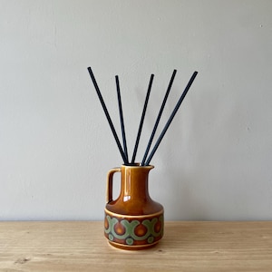 May include: A brown ceramic vase with a handle, containing black reeds. The vase features a decorative pattern of green and orange shapes. It rests on a wooden surface against a neutral wall.