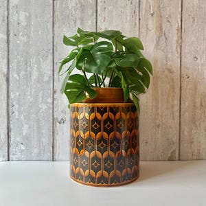 May include: A cylindrical ceramic planter featuring a glossy finish with a repeating geometric pattern in shades of dark brown and amber. The planter contains a green artificial monstera plant with multiple split leaves extending upwards.