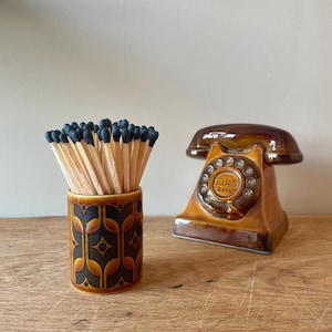 May include: A ceramic match holder with a geometric pattern and a vintage telephone bank. The match holder is filled with matches with black tips. The telephone bank is brown and has the words "TELEPHONE BANK" on the dial.
