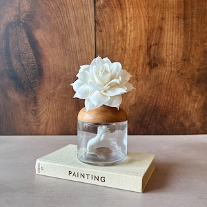 May include: A clear glass jar with a rounded wooden lid topped by a white flower-shaped diffuser. A white cotton wick extends from the flower into the jar. The jar rests on a cream-coloured book with the word PAINTING and the number 26 printed on it.