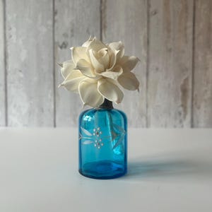 May include: A small, cylindrical, translucent blue glass bottle featuring a delicate white and gold painted floral motif around its centre, topped with a large, cream-coloured, multi-layered wooden flower bloom that sits securely in the bottle neck.