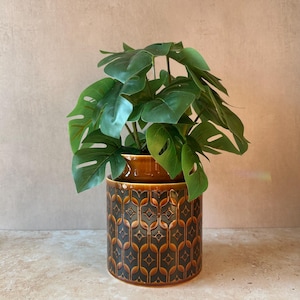 May include: A cylindrical ceramic planter featuring a dark brown and black geometric pattern with a glossy finish. The planter holds a lush, artificial monstera plant with broad, split green leaves that extend outward from the centre.