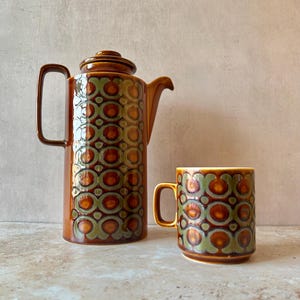 May include: A tall ceramic coffee pot with a lid and a matching mug, both featuring a repeating geometric pattern in shades of brown and olive green. The coffee pot has a side handle and a spout, while the mug has a single handle on the side.