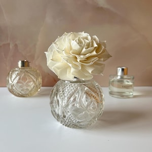 May include: A clear, round, cut-glass bottle with a faceted diamond pattern, topped with a large, cream-coloured, layered wooden flower diffuser. Two additional glass perfume bottles are positioned in the background, one textured and one smooth.