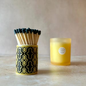 May include: A cylindrical ceramic match holder featuring a black and yellow geometric quatrefoil pattern, filled with wooden matches that have dark green tips. Beside it sits a frosted yellow glass candle with a circular white label.