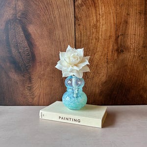 May include: A small, light blue, speckled glass vase with a bulbous, hourglass shape. A single, large, off-white flower with layered petals sits inside the vase. The vase rests on a cream-coloured book with the word PAINTING printed on the spine.