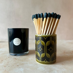 May include: A black glass candle holder with a circular white label and a cylindrical ceramic match holder featuring a dark green and black geometric pattern filled with wooden matches that have black tips.