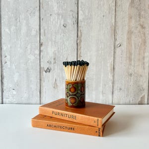 May include: A cylindrical ceramic match holder with a brown and green geometric pattern, filled with wooden matches with black tips, resting on top of two stacked tan hardcover books labelled Furniture and Architecture on their spines.