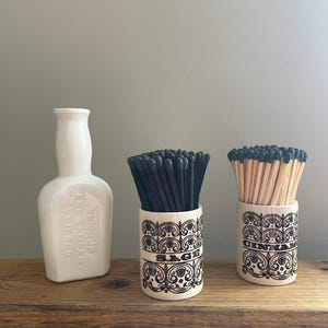 May include: Three ceramic containers on a wooden surface. A white bottle-shaped container with text, and two cylindrical containers with black matches and wooden matches. The cylindrical containers have a black and white floral design and the words "Sage" and "Cinnamon".