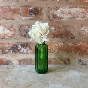 May include: A small, rectangular, emerald green glass bottle with vertical ribbed sides and embossed text on the front, containing a single, multi-layered, off-white wooden flower with pointed petals arranged in a blooming, circular pattern.