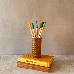 May include: A cylindrical brown ceramic pencil holder with a textured honeycomb pattern, containing six coloured pens. The holder rests on two stacked hardcover books, one yellow with the text COTSWOLDS and one brown with the text ARCHITECTURE.