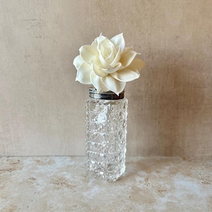 May include: A clear, square-shaped glass bottle with a textured, diamond-cut pattern on its sides. The bottle features a metallic silver-toned cap and is topped with a single, large, off-white decorative flower made of thin, layered petals.