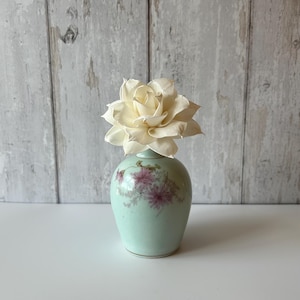 May include: A small, light mint green ceramic vase with a rounded body and a narrow neck, featuring a delicate hand-painted floral design in shades of pink and green. A single, large, cream-coloured artificial flower sits inside the vase opening.