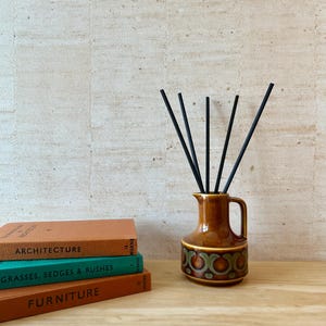 May include: A brown ceramic jug with a handle and a repeating green and brown circular pattern around the base, containing five thin black reed diffuser sticks. Next to the jug are three stacked books labelled Architecture, Grasses, Sedges & Rushes, and Furniture.