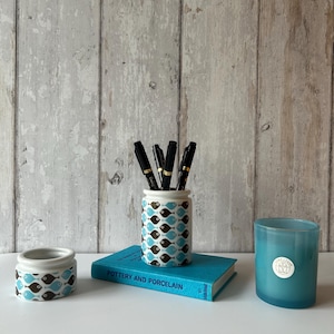 May include: A ceramic pen holder with a repeating blue and brown fish pattern, a matching small ceramic bowl, a turquoise book titled Pottery and Porcelain, and a cylindrical turquoise candle with a white circular label on the front.