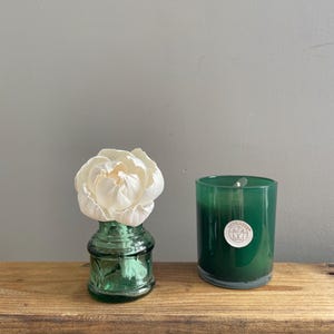 May include: A green glass vase with a white flower and a green candle in a glass jar sit on a wooden surface. The candle has a white label with a circular design. The items are arranged against a grey wall.