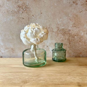 May include: A clear, light green glass bottle with a short neck containing a large, white, multi-petaled flower on a stem, positioned next to a smaller, empty, matching light green glass bottle with a wide, cylindrical neck and a thick rim.