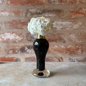 May include: A small, dark brown glass bud vase with a slender neck and a wide, clear glass base. The vase features a metallic gold-coloured collar at the top and holds a single, large, white artificial flower with layered, ruffled petals.