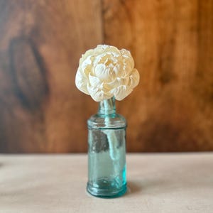 May include: A single cream-coloured flower with layered, delicate petals sits inside a small, translucent teal-tinted glass bottle. The bottle has a cylindrical body with a narrow neck and a slightly flared rim, holding the stem of the flower upright.