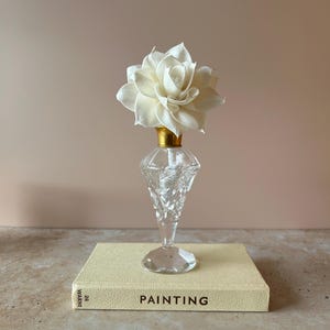 May include: A clear, faceted glass perfume bottle with a gold-toned neck, topped with a large, cream-coloured fabric flower. The bottle rests on a light yellow book with the word PAINTING and the number 26 printed on the spine.