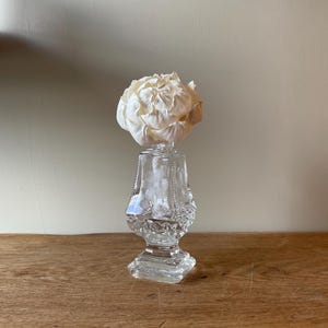 May include: A clear glass perfume bottle with a square base and a faceted body featuring etched floral patterns. A large, white, layered paper flower is inserted into the top opening of the bottle, serving as a decorative diffuser element.