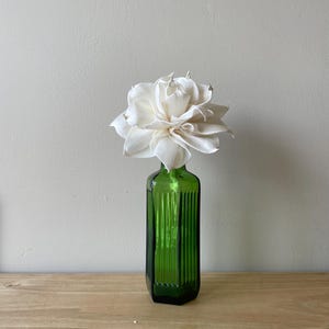 May include: A single cream-coloured sola wood flower with layered, pointed petals sits inside a tall, rectangular, emerald green glass bottle. The bottle features vertical ribbed detailing along its four sides and a narrow neck holding the flower.