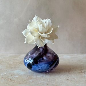 May include: A small, rounded glass vase featuring a swirling blend of deep purple, blue, and black colours. A single, multi-layered cream-coloured flower with pointed petals is positioned in the narrow opening at the top of the glass vessel.