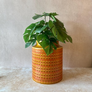 May include: A cylindrical ceramic vase featuring a repeating orange and yellow floral pattern with a smooth finish. The vase contains a lush green artificial plant with broad, split leaves that extend outward from the top opening of the vessel.
