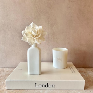 May include: A white ceramic diffuser bottle topped with a large, cream-coloured sola wood flower, placed next to a white candle in a glass jar. Both items sit on a thick, rectangular white book with the word London printed on the front cover.